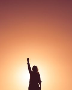 Silouhette of long-haired individual holding fist up in front of sunset