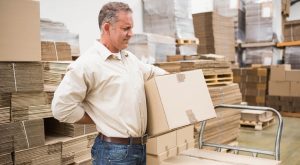 Man in warehouse holding cardboard box and lower back in discomfort