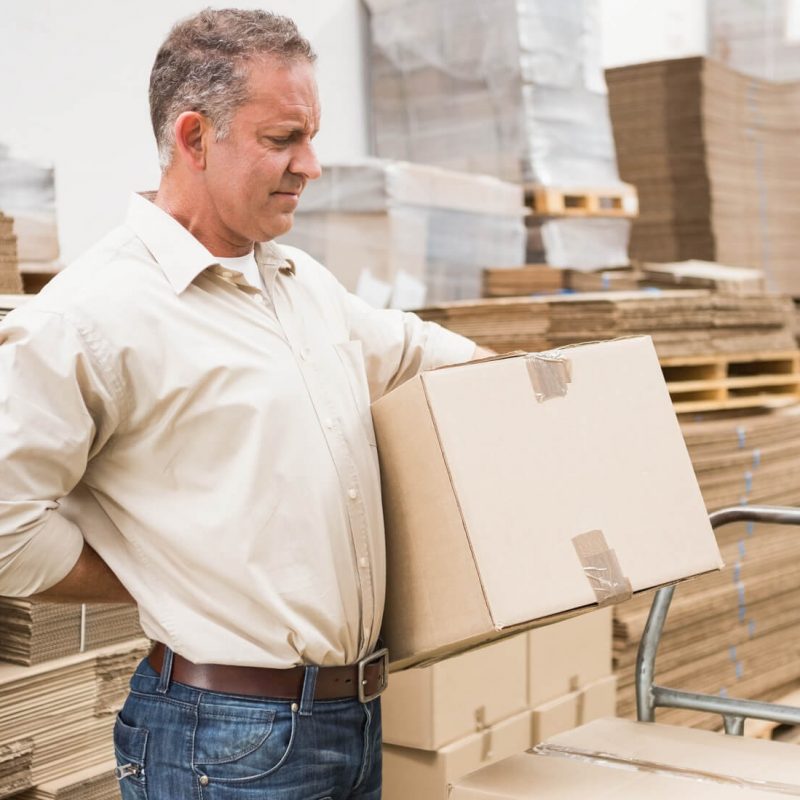 Man in warehouse holding cardboard box and lower back in discomfort