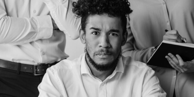 Confused man seated in front of standing woman writing in a small book and man with folded arms